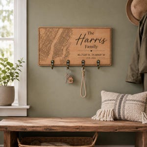 May include: A rectangular wooden wall-mounted key holder featuring a black engraved map on the left side. The right side displays the text The Harris Family, coordinates 40.7128 N, 74.0060 W, and EST. 2024. It includes four bronze-colored metal hooks.