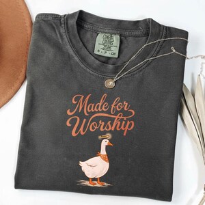 Puede incluir: Camiseta gris oscuro con el texto "Made for Worship" en escritura naranja. La camiseta presenta un dibujo de un ganso con un halo y un pañuelo. Un collar plateado con un colgante está sobre la camiseta.