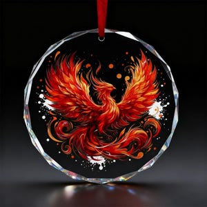 Puede incluir: Adorno de cristal facetado redondo con un vibrante diseño de fénix. El fénix está representado en tonos rojos y naranjas sobre un fondo negro, con detalles blancos. Se adjunta una cinta roja para colgar.