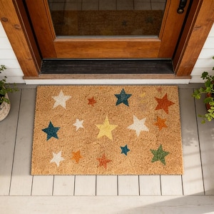 May include: A rectangular doormat made of natural coir fibre. The mat features a repeating pattern of colourful stars in various sizes and colours, including white, yellow, green, orange, and blue. The mat is placed in front of a wooden door.