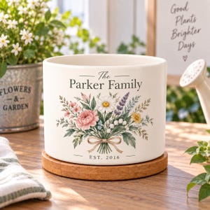 May include: A white cylindrical ceramic planter resting on a circular wooden base. The front features a colourful floral bouquet illustration with a ribbon, the text "The Parker Family" above, and "EST. 2016" printed below the flowers.