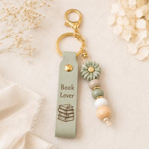 May include: A sage green leather keychain featuring a gold-tone metal clasp and ring. The strap is embossed with the text Book Lover and a stack of books illustration. A decorative strand of beads in sage, beige, white, and wood tones hangs beside it.