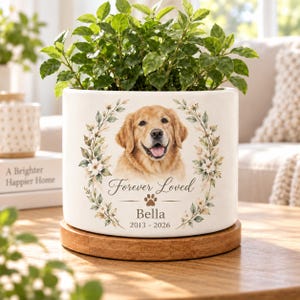 May include: A white ceramic cylindrical planter on a round wooden base containing a green plant. The front features a golden retriever illustration framed by floral wreaths, with the text Forever Loved, Bella, and the years 2013-2026.