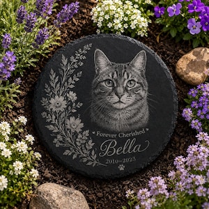 May include: A round black slate memorial stone featuring an engraved portrait of a tabby cat. To the left is a floral vine design. The text reads Forever Cherished, Bella, and 2010-2023, with a small paw print icon below the dates.