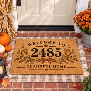 May include: A rectangular tan coir doormat featuring the black text WELCOME TO 2485 THANKFUL HOME. The design includes illustrations of golden wheat stalks, a small red maple leaf, a red ribbon bow, and a small red heart centred below the text.