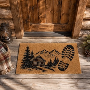 May include: A rectangular doormat made of natural fibers. The mat features a black graphic of a cabin, mountains, and trees, with a large boot print. The mat is placed in front of a wooden door.