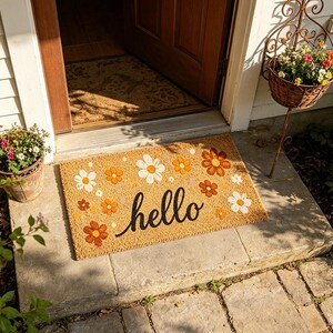 Puede incluir: Una alfombra rectangular con un fondo beige y un diseño floral en blanco y naranja. La palabra "hello" está escrita en negro en el centro. La alfombra está colocada en un escalón de piedra frente a una puerta de madera.