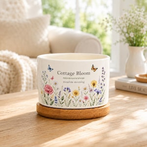 May include: A cylindrical white ceramic bowl featuring a colourful botanical print of wildflowers and butterflies. The text Cottage Bloom and meadow morning is printed on the front. The bowl rests on a circular light brown wooden base.