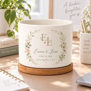 Könnte beinhalten: Ein zylindrischer Übertopf aus weißer Keramik auf einem runden Holzsockel. Die Vorderseite zeigt ein florales Kranzmotiv in Grün und Rosa, das die goldenen Initialen EL, die Namen Emma & Liam, das Datum JUNE 14, 2026 und den Text grown together in love umrahmt.