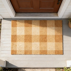 Può includere: Un zerbino rettangolare con un motivo a quadri nei toni del beige e marrone chiaro. Il tappeto è realizzato in un materiale strutturato ed è posto su un portico in legno davanti a una porta marrone.