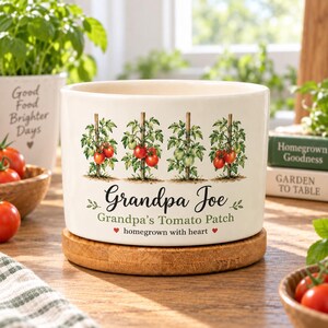 Puede incluir: Una maceta de cerámica blanca sobre una base de madera redonda. La maceta presenta un gráfico de cuatro plantas de tomate en estacas, con el texto "Grandpa Joe", "Grandpa's Tomato Patch" y "homegrown with heart" impreso en negro y verde.