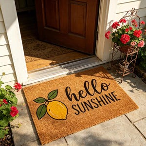 Puede incluir: Felpudo rectangular de fibra natural, con una ilustración de limón amarillo con hojas verdes y la frase "hello SUNSHINE" en negro. La alfombra está colocada frente a una puerta de madera.