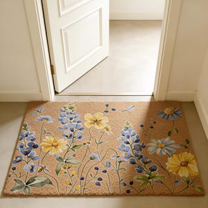 May include: A rectangular doormat with a floral design featuring blue, yellow, and green flowers. The mat is made of a natural fiber material and is placed in front of an open white door. The flowers are painted in a watercolor style.