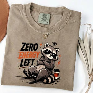 May include: A tan Comfort Colors t-shirt featuring a graphic of a raccoon wearing sunglasses with the text "ZERO ENERGY LEFT". The raccoon is sitting with its arms crossed, next to a coffee cup. A silver necklace is draped across the shirt.