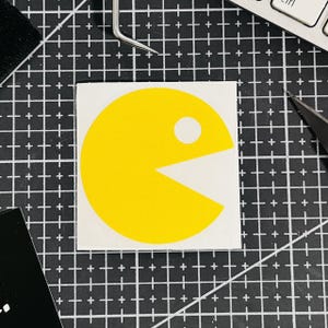 Könnte beinhalten: Ein gelber Pac-Man-Aufkleber mit weißem Hintergrund. Die klassische Videospiel-Figur ist mit offenem Mund und einem weißen Punkt dargestellt. Der quadratische Aufkleber liegt auf einem schwarz-weißen Raster.