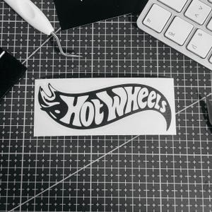 May include: A black and white Hot Wheels logo sticker. The logo features the words "Hot Wheels" in a stylized font, with a flame design. The sticker is on a cutting mat with a keyboard and tools in the background.