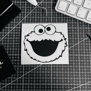 Könnte beinhalten: Ein schwarz-weißer Aufkleber mit dem Gesicht des Krümelmonsters. Der Aufkleber hat einen großen offenen Mund, runde Augen und eine pelzige Kontur. Der Aufkleber befindet sich auf einem weißen Quadrat, umgeben von einer schwarzen Schneidematte, einer Tastatur und Werkzeugen.