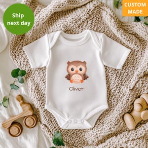 May include: White baby onesie with a brown owl illustration and the name "Oliver" printed below. The owl has large orange eyes and is centered on the front of the onesie. The words "Custom Made" are in the top right corner.