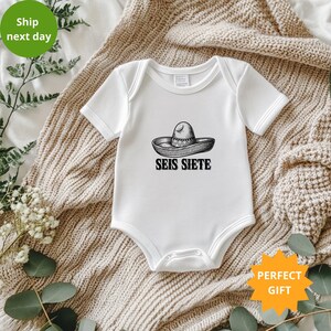 May include: White baby bodysuit featuring a sombrero illustration and the words "SEIS SIETE" in black. The short-sleeved bodysuit has a snap closure. The background is a beige blanket.