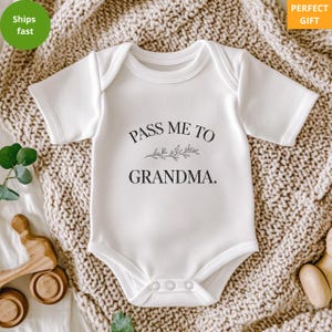 May include: White baby onesie with the text "PASS ME TO GRANDMA." in black, with a sprig design. The onesie has short sleeves and snaps at the bottom. The background is a beige knitted blanket.