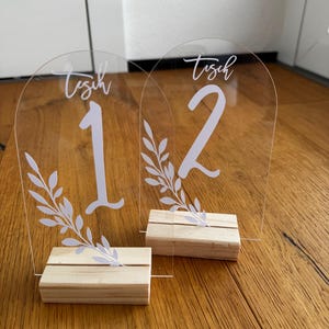 Può includere: Due numeri da tavolo in acrilico trasparente, 1 e 2, con scritte bianche e decorazioni a foglie. Ogni numero è posizionato su una piccola base di legno. La parola "Tisch" è scritta sopra ogni numero.