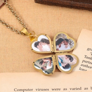 May include: A gold-colored, four-leaf clover locket necklace with heart-shaped compartments, each holding a photograph. The necklace is on a chain and rests on a tan surface. The locket is open, revealing the photos.