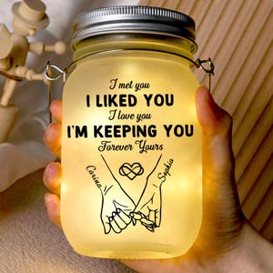 Puede incluir: Un tarro Mason brillante con tapa y asa plateadas. El tarro tiene el texto "I met you, I liked you, I love you, I'm keeping you, Forever Yours." Debajo del texto hay un gráfico de dos manos tomadas. Los nombres "Carina" y "Sophia" están escritos.