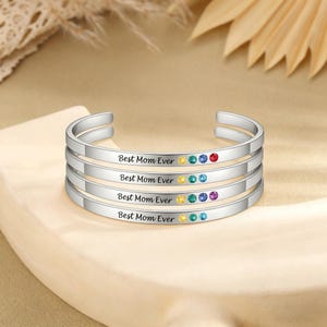 Puede incluir: Brazaletes de plata, cada uno grabado con "Best Mom Ever" y adornado con piedras preciosas de colores. Los brazaletes están apilados, mostrando una variedad de colores de gemas, incluyendo rojo, azul, verde, amarillo y morado.