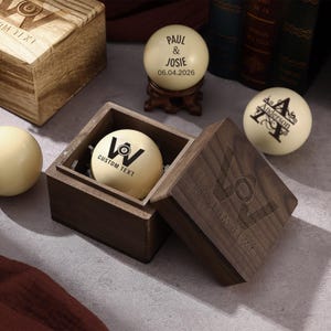 May include: A selection of personalised billiard balls and wooden boxes. One ball displays "PAUL & JOSIE" and the date "06.04.2026". Another ball has the name "Anderson". The boxes are engraved with a logo and the text "CUSTOM TEXT".