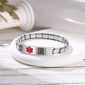 Puede incluir: Pulsera de identificación médica de acero inoxidable con un símbolo médico rojo y un código QR. La pulsera tiene las palabras "HEALTH INFO" y "SCAN ME". La pulsera está sobre una superficie blanca.