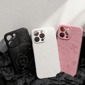 May include: Three phone cases in black, white, and pink. Each case features a decorative embossed pattern with a cross and circular designs. The cases are made of a smooth material and are designed to fit a smartphone.