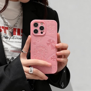 Puede incluir: Funda de teléfono rosa con cruces y diseños circulares en relieve. La funda tiene un recorte para la cámara y se sostiene en una mano. La funda tiene un diseño decorativo.