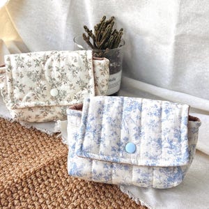 May include: Two small quilted pouches with flap closures and snap buttons. One pouch features a cream fabric with a brown floral print and a white snap, while the other has a cream fabric with a blue floral print and a light blue snap button.