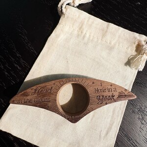 May include: A wooden book page holder with the text "Head in the Clouds" and "Nose in a Book" engraved on it. The holder is brown and has a circular opening. It rests on a beige drawstring bag.