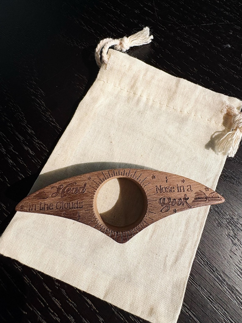 May include: A wooden book page holder with the text "Head in the Clouds" and "Nose in a Book" engraved on it. The holder is brown and has a circular opening. It rests on a beige drawstring bag.