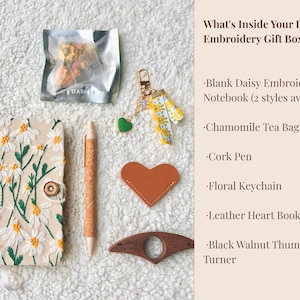 May include: An overhead shot of a gift box's contents, including a daisy-embroidered notebook, chamomile tea bag, cork pen, floral keychain, leather heart bookmark, and a black walnut thumb page turner. The text reads "What's Inside Your Daisy Embroidery Gift Box:"