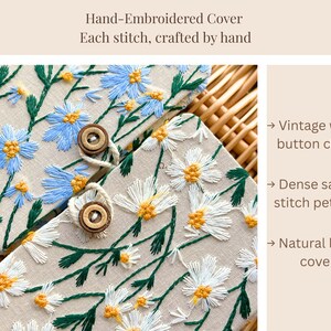 May include: Hand-embroidered cover with blue and white daisy floral design. Features a vintage wooden button closure and dense satin stitch petals. The text reads: Hand-Embroidered Cover. Each stitch, crafted by hand.