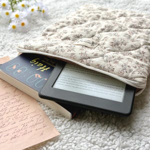 May include: A quilted, floral-patterned e-reader case in cream and brown, with a zipper closure. The case holds a black e-reader and a book titled "Haig". The book has a blue cover with white text and illustrations.