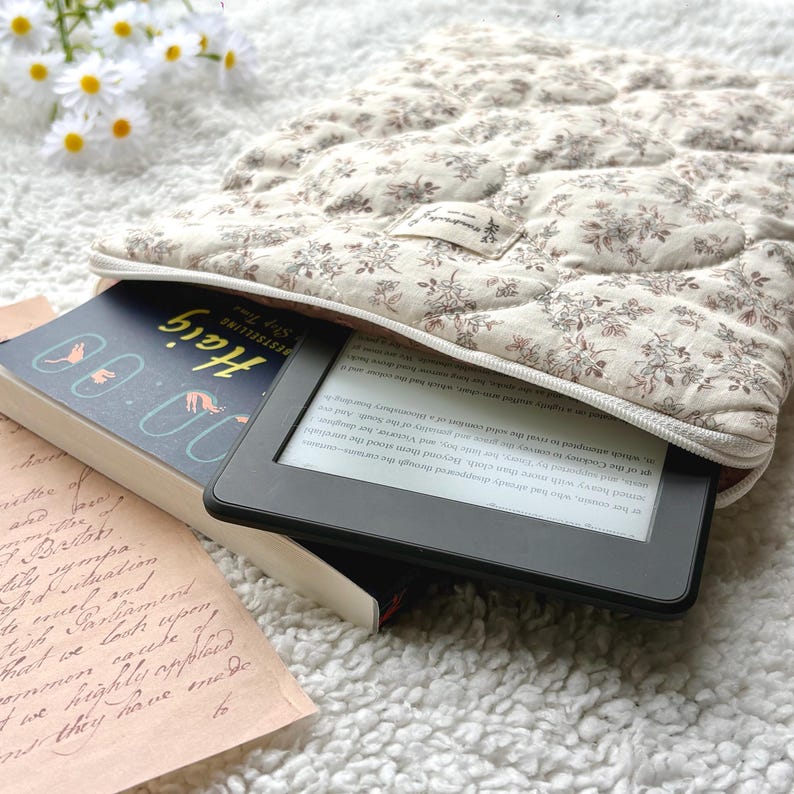 May include: A quilted, floral-patterned e-reader case in cream and brown, with a zipper closure. The case holds a black e-reader and a book titled "Haig". The book has a blue cover with white text and illustrations.