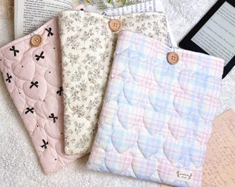 May include: Three quilted book covers in soft pastel colors. One is pink with black bow accents, another has a floral pattern, and the third features a heart-shaped pattern in blue and pink. Each cover has a wooden button closure.