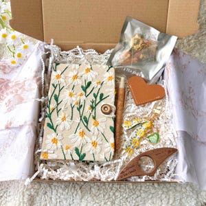 May include: A gift box filled with a daisy-embroidered journal, a pen, a leather heart, a keychain, a wooden bookmark, and a tea bag. The journal has a button closure. The items are arranged on white paper shreds.