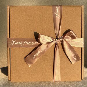May include: A brown cardboard gift box tied with a light brown satin ribbon. The ribbon is wrapped around the box and tied in a bow. The words "Just for you" are printed on the ribbon in white.