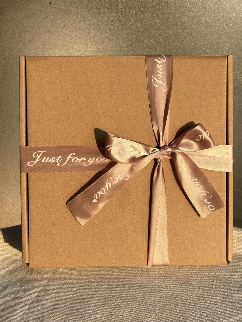 May include: A brown cardboard gift box tied with a light brown satin ribbon. The ribbon is wrapped around the box and tied in a bow. The words "Just for you" are printed on the ribbon in white.