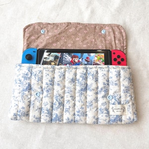 May include: A quilted case for a Nintendo Switch console. The case features a blue and white floral pattern on the front and a brown floral pattern on the inside flap. The console is visible, displaying a video game.