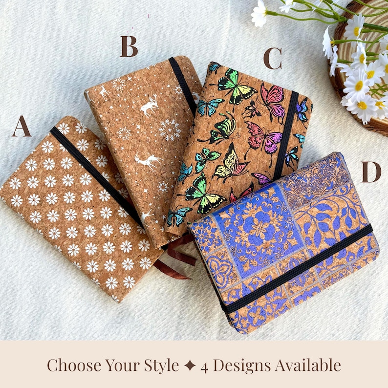 May include: Four cork notebooks with different designs: white daisies, deer and snowflakes, colorful butterflies, and blue floral patterns. Each notebook has a black elastic band and a ribbon bookmark. The text "Choose Your Style 4 Designs Available" is at the bottom.