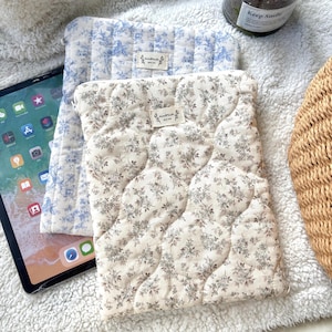 May include: Two rectangular quilted tablet sleeves featuring floral patterns. One sleeve has a blue and white toile print, while the other has a beige and brown floral design. Both sleeves include a small rectangular label reading Handmade with love.