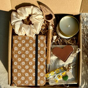 May include: A gift box filled with various items. Includes a cream-colored scrunchie, a cork notebook with a daisy pattern, a pen, a candle, a leather heart, a keychain, and a wooden bookmark. The box is brown.