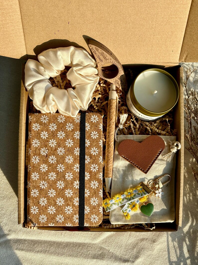 May include: A gift box filled with various items. Includes a cream-colored scrunchie, a cork notebook with a daisy pattern, a pen, a candle, a leather heart, a keychain, and a wooden bookmark. The box is brown.