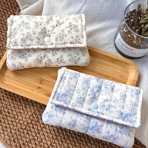 May include: Two rectangular quilted pouches with flap closures. The top pouch features a cream fabric with a small-scale brown and green floral print and a white snap button. The bottom pouch has a white fabric with a blue floral pattern and a light blue snap button.
