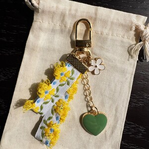 May include: A floral keychain with a gold clasp and chain. The keychain features a white ribbon with yellow and blue flower embroidery, yellow fringe, a white flower charm, and a green heart charm. The keychain rests on a beige drawstring bag.